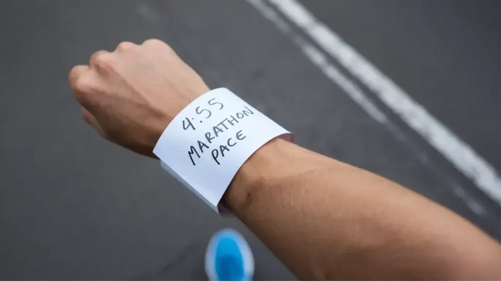 a piece of paper that has been wrapped around the wrist of a runner. the paper is blank, white, but has the words "4:55 Marathon Pace" written on it in pen. the image is POV of the runner.