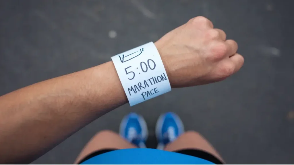 a piece of paper that has been wrapped around the wrist of a runner. the paper is blank, white, but has the words "5 hour Marathon Pace" written on it in pen. the image is POV of the runner.