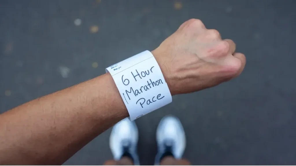 a piece of paper that has been wrapped around the wrist of a runner. the paper is blank, white, but has the words "6 hour Marathon Pace" written on it in pen. the image is POV of the runner.