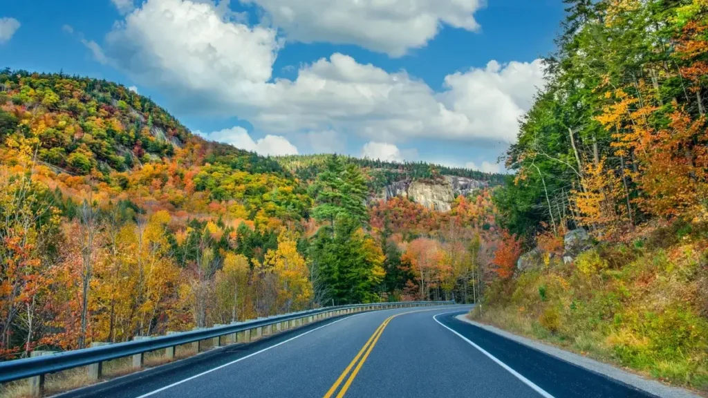 A two-lane road curves through colorful fall foliage and forested hills in New Hampshire, with a rocky cliff visible in the distance under a partly cloudy sky.
