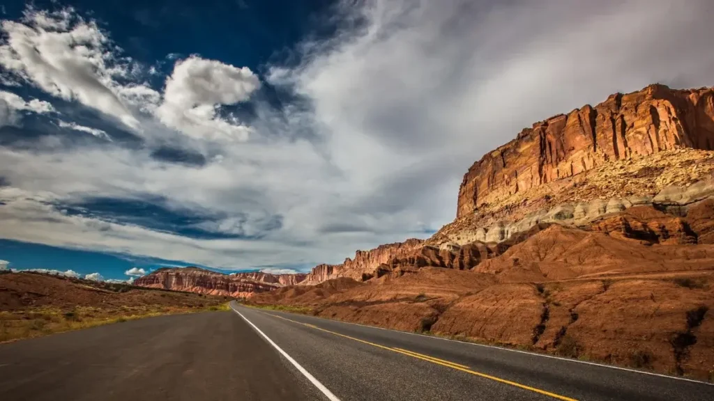 Open road winding past dramatic red rock cliffs under a partly cloudy sky in Utah.